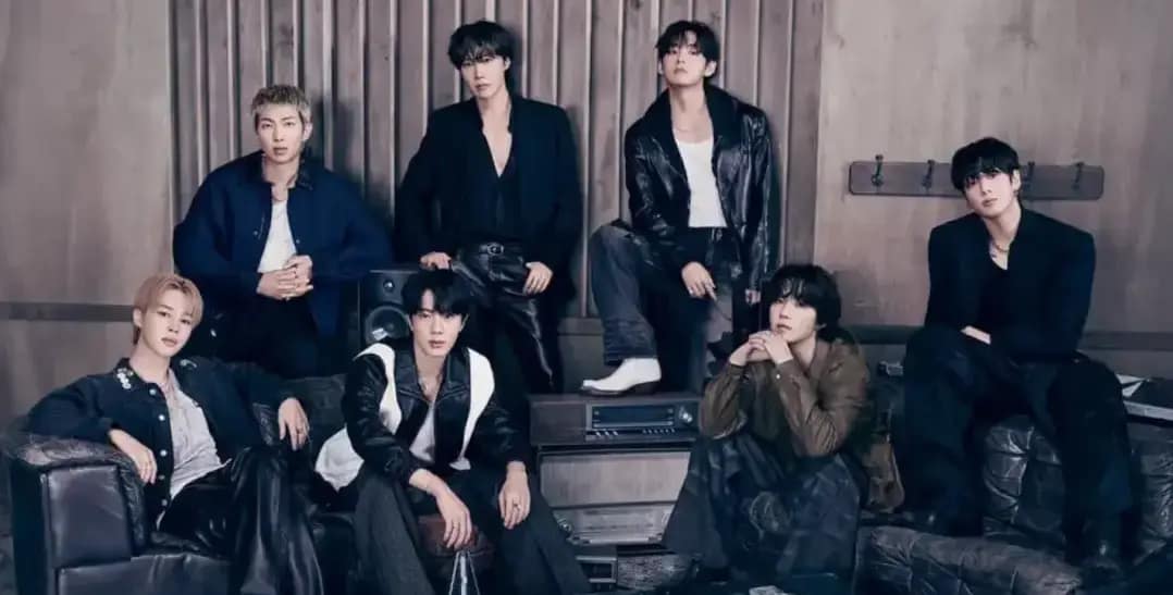 Integrantes do BTS posam juntos em um ambiente interno, usando roupas escuras em uma fotografia promocional do grupo sul coreano