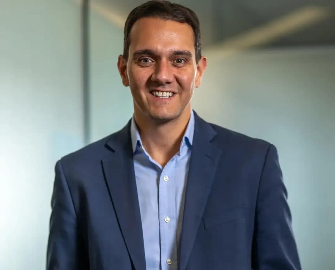 Rodolfo Santos posa para retrato corporativo usando blazer azul-marinho e camisa azul-clara, com as mãos nos bolsos, diante de um fundo claro