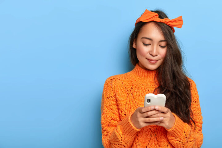 Photo of lovely millennial girl uses mobile phone, creats own internet web page, focused in screen, wears loose orange jumper, headband tied in bow, poses over blue background, free space aside