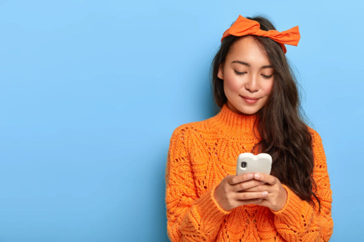 Photo of lovely millennial girl uses mobile phone, creats own internet web page, focused in screen, wears loose orange jumper, headband tied in bow, poses over blue background, free space aside Photo of lovely millennial girl uses mobile phone, creats own internet web page, focused in screen, wears loose orange jumper, headband tied in bow, poses over blue background, free space aside