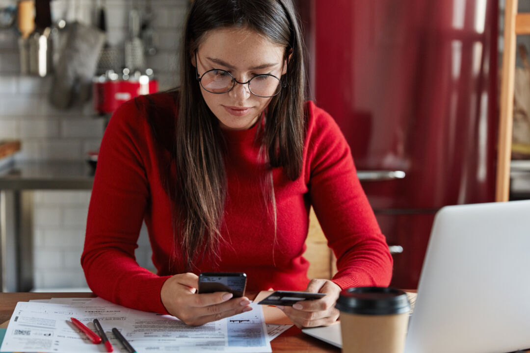 Cropped image of brunette freckled woman in red casual clothes, holds modern smart phone and plastic card in hands, checks bank account, works with financial documents, drinks coffee, poses in kitchen