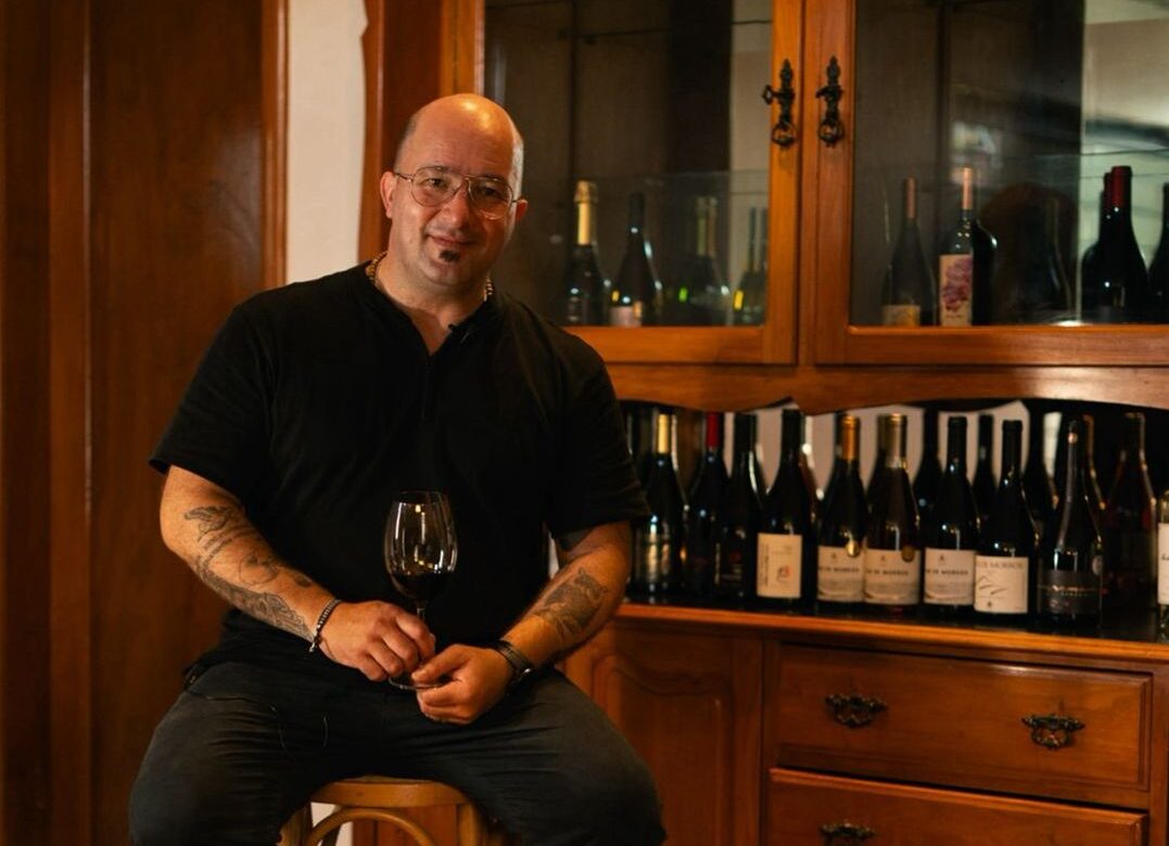 Sommelier Gustavo Giacchero segura uma taça de vinho durante degustação, sentado em frente a uma adega com garrafas variadas, em ambiente aconchegante de madeira.