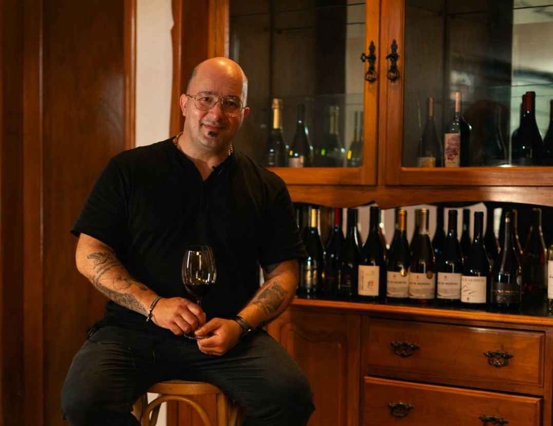 WhatsApp Image 2025 10 27 at 18 21 43 Sommelier Gustavo Giacchero segura uma taça de vinho durante degustação, sentado em frente a uma adega com garrafas variadas, em ambiente aconchegante de madeira.