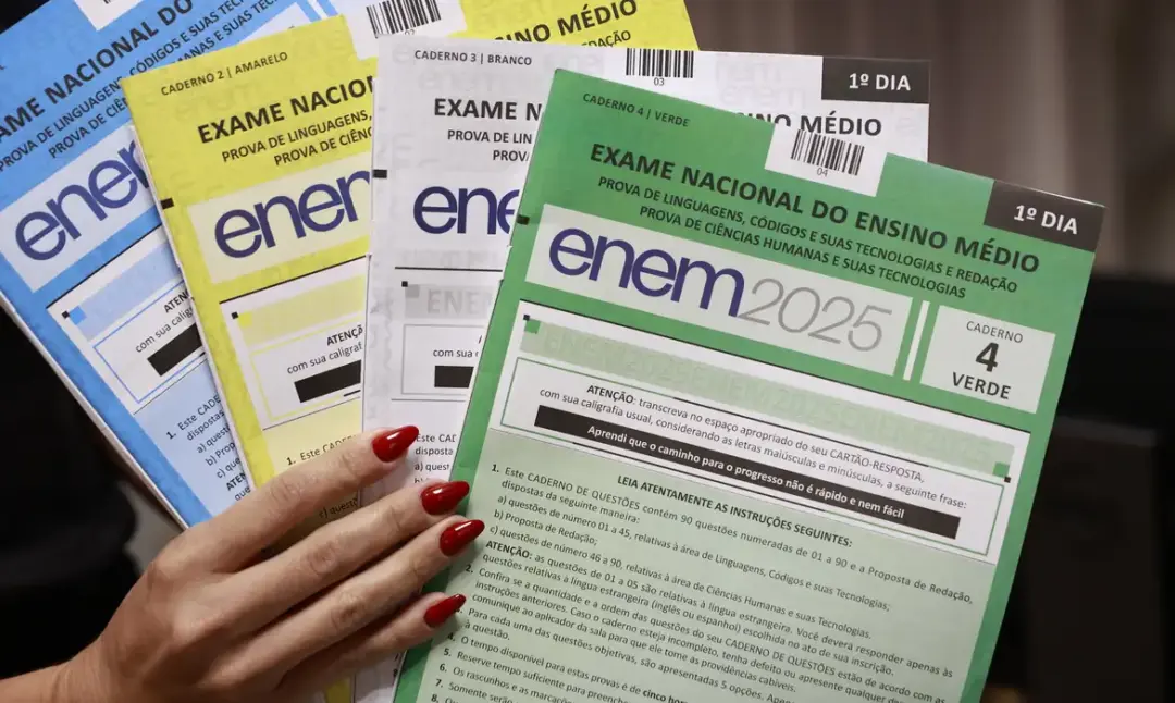 Uma pessoa segurando diferentes cadernos de prova do Enem 2025, em cores variadas, representando os modelos aplicados no exame