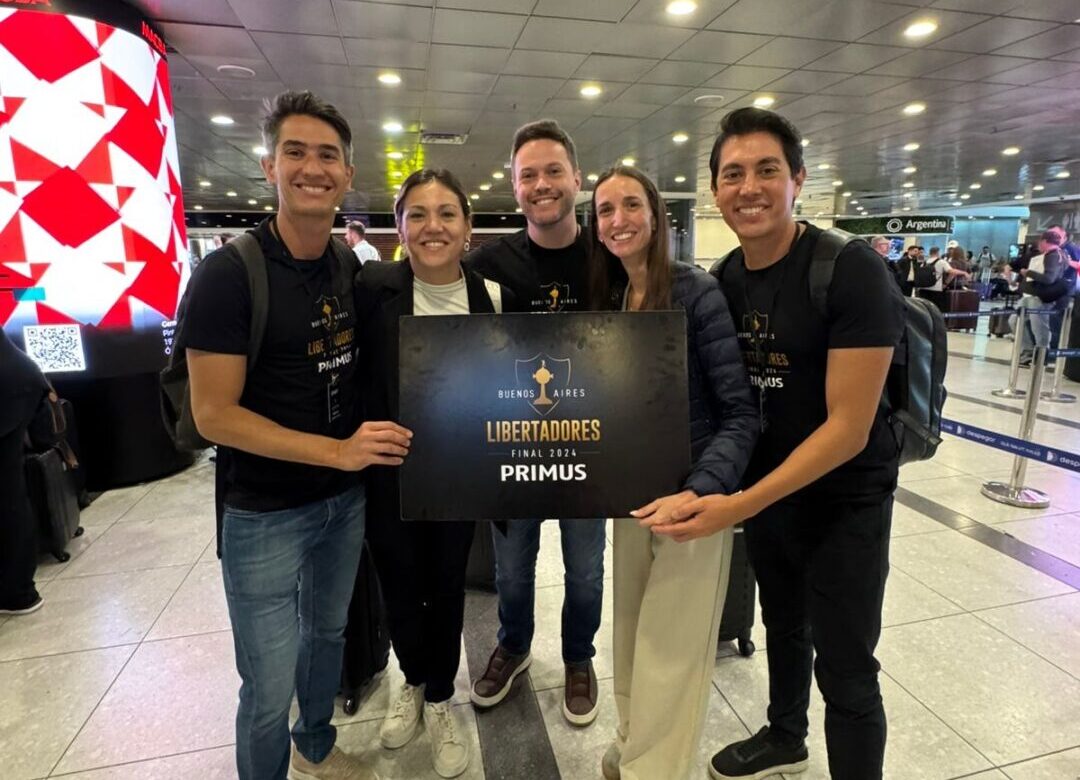 Cinco integrantes da Primus Turismo posam sorridentes em aeroporto segurando placa da final da Libertadores 2024 em Buenos Aires.