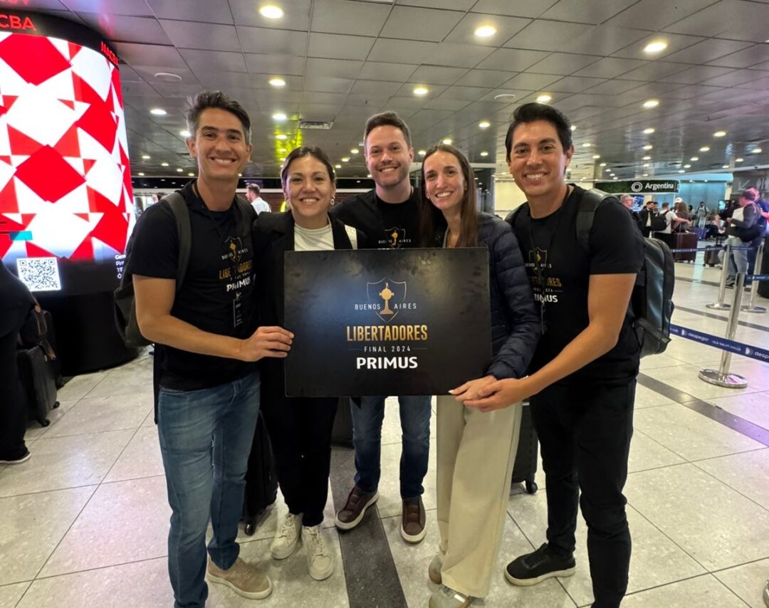 WhatsApp Image 2025 11 01 at 10 24 56 Cinco integrantes da Primus Turismo posam sorridentes em aeroporto segurando placa da final da Libertadores 2024 em Buenos Aires.