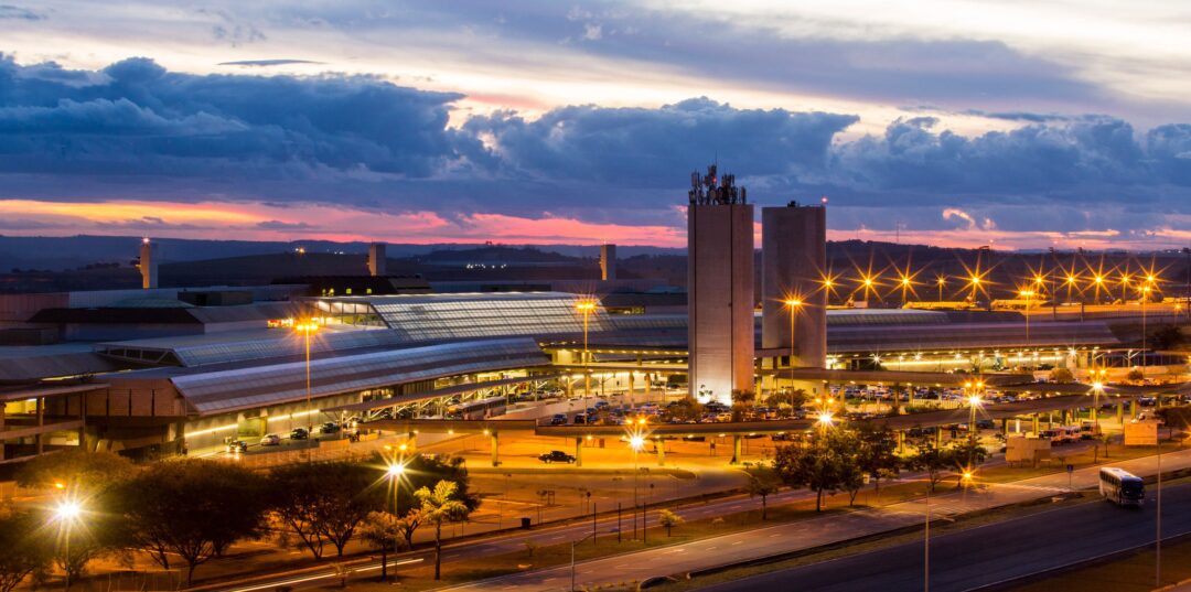Fachada do BH Airport iluminado ao entardecer, com movimento de veículos e estrutura do terminal em destaque