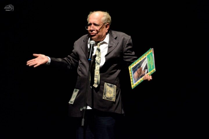 Saulo Laranjeira se apresenta no palco com microfone e figurino simbólico, unindo humor e crítica social em espetáculo Saulo Laranjeira em cena mistura humor e crítica social em espetáculo