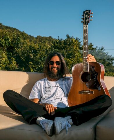 Armandinho retorna a BH com repertório que marcou o reggae brasileiro Armandinho aparece sentado em sofá ao ar livre segurando um violão acústico. O cantor usa camiseta branca, óculos escuros e sorri para a câmera com paisagem verde ao fundo