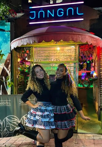 Arraiá do Jângal leva clima junino para a noite de BH com música e tradição Duas mulheres com trajes típicos de festa junina posam sorrindo em frente à entrada iluminada do Jângal, em Belo Horizonte, durante evento temático junino