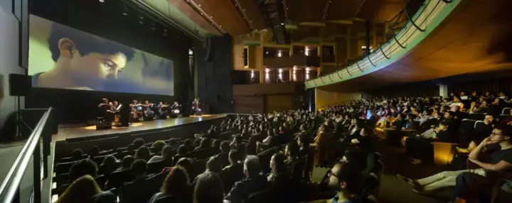 Público ocupa auditório durante atividade audiovisual do CineBH, com músicos no palco e imagens projetadas em uma grande tela; Ana Lúcia Torre 