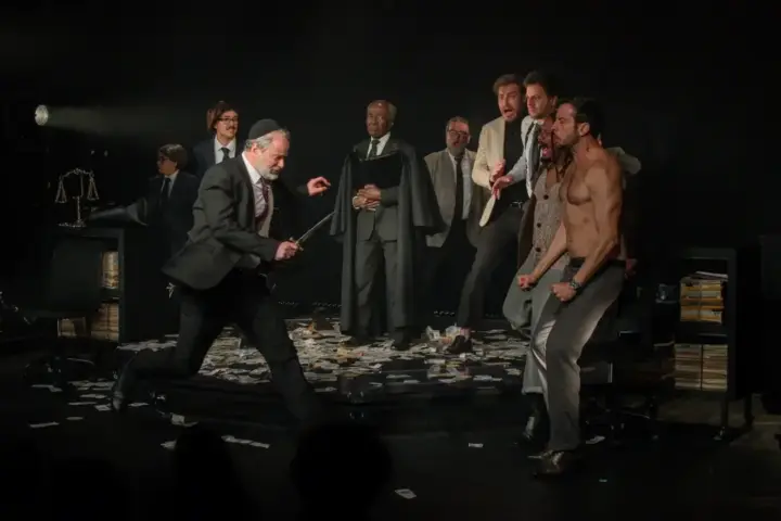 Igor Dallegrave 6 Atores encenam uma cena de “O Mercador de Veneza” em palco de teatro, com figurinos contemporâneos e cenário que remete a um julgamento
