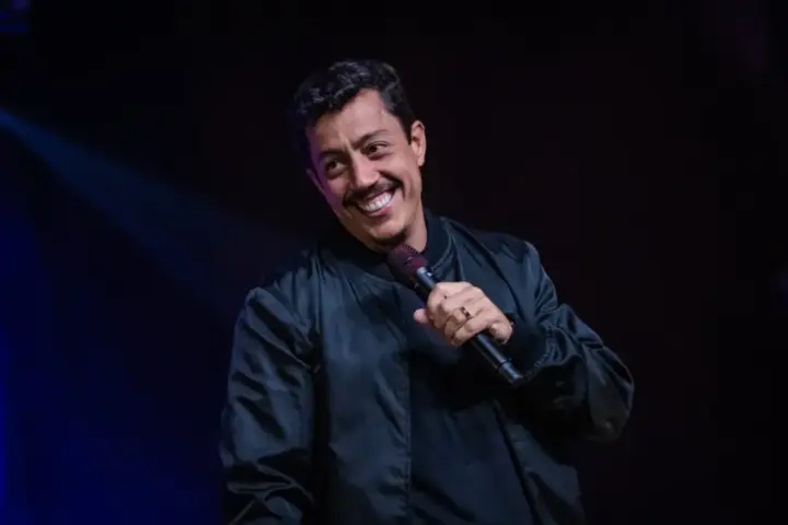 Renato Albani segura um microfone e sorri durante apresentação de stand-up em palco