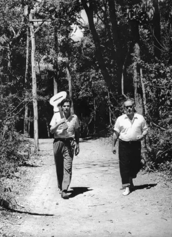 “Brasília: Sinfonia da Alvorada” homenageia JK em apresentação em Belo Horizonte Fotografia em preto e branco mostra dois homens caminhando por uma área arborizada, em imagem relacionada à obra “Brasília: Sinfonia da Alvorada”