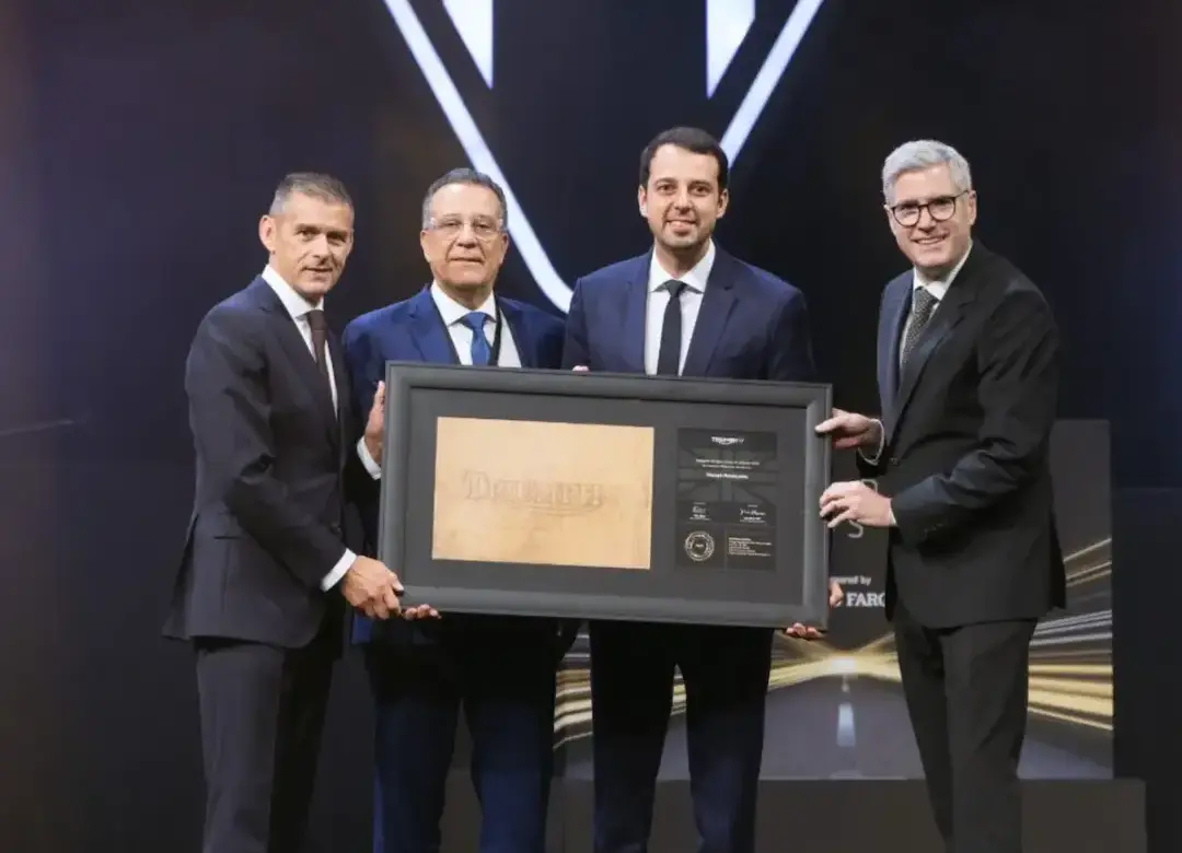 Representantes da Triumph BHR posam em um palco durante uma cerimônia de premiação, segurando juntos um quadro comemorativo
