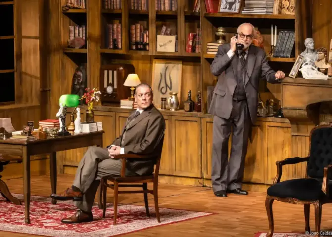 Cena do espetáculo A Última Sessão de Freud, com dois atores interpretando Sigmund Freud e C. S. Lewis em um escritório repleto de livros, móveis de madeira e objetos decorativos
