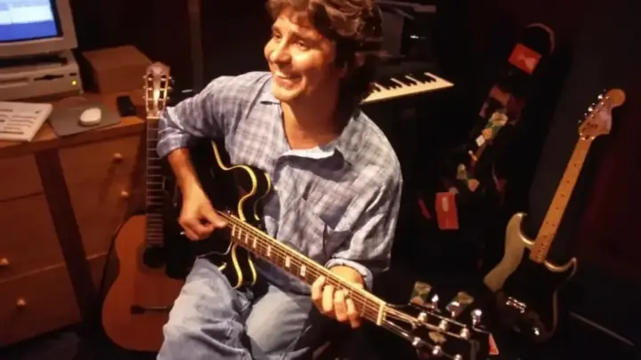 Lô Borges aparece sorrindo enquanto toca guitarra em um estúdio musical.